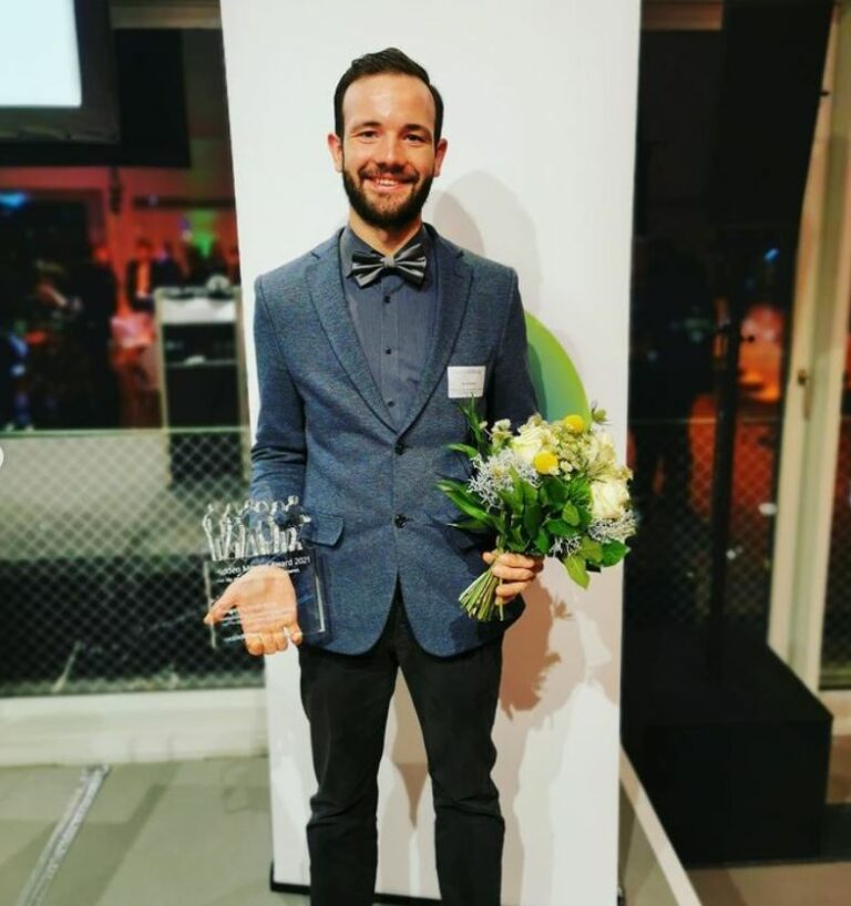 Mann mit dunklem Haar im blauem Anzug mit Blumenstrauß und Trophäe lächelt