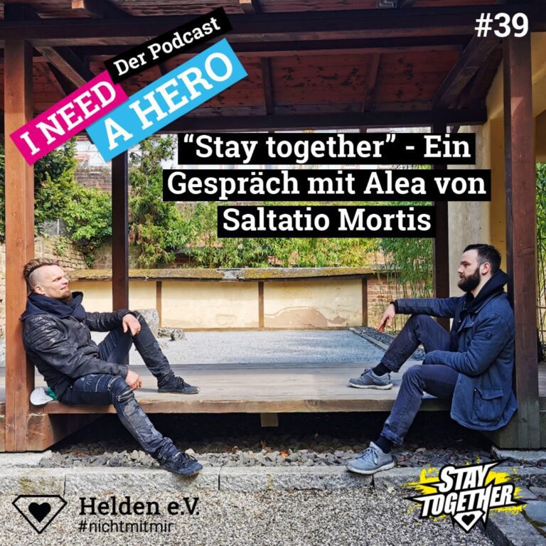 Zwei Männer sitzen in Holzpavillion. Mittig Schriftzug "Ein Gespräch mit Alea von Saltatio Mortis". Links oben Schriftzug in verschiedenen Farben: "I need a Hero - Der Podcast"