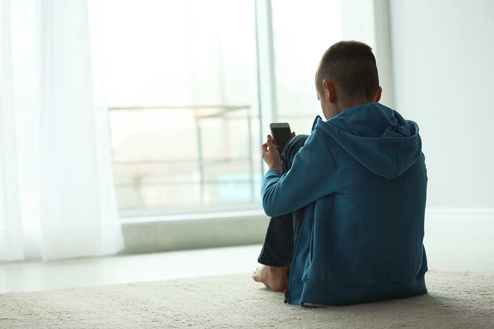Junge in blauem Pullover schaut zusammengekauert auf dem Boden sitzendend auf ein Smartphone. Rückansicht.
