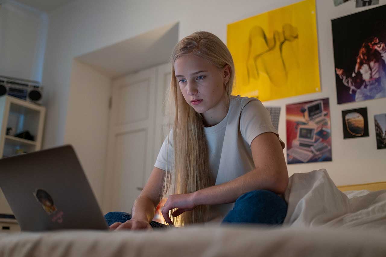 Blondes Mädchen in ihrem Kinderzimmer auf ihrem Bett im Schneidersitz sitzend schaut ausdruckslos auf ihren Laptop der auf dem Bett steht.