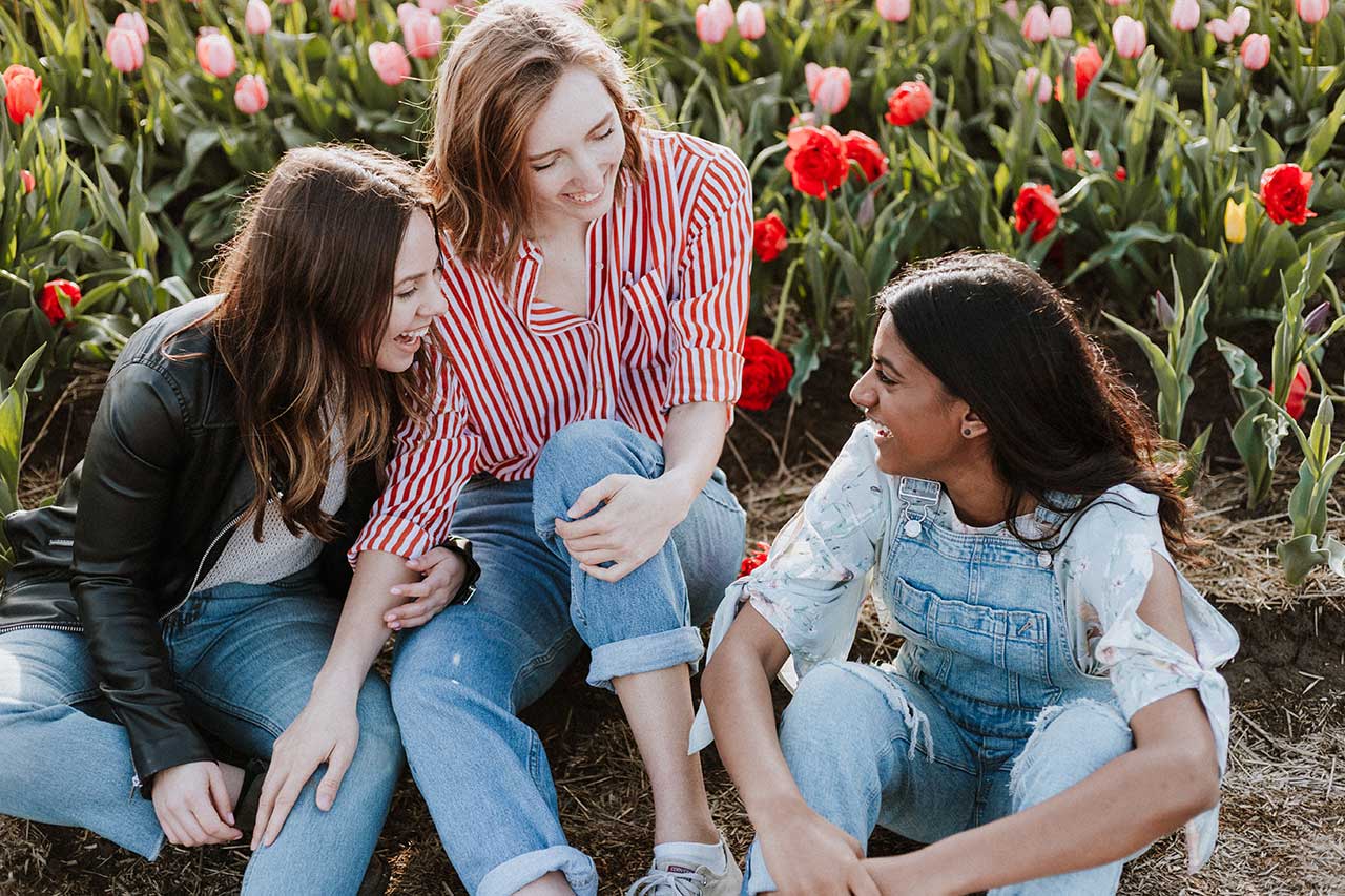Drei junge Frauen amüsieren sich gemeinsam in einem Blumenbeet sitzend.
