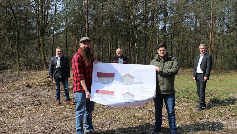 Zwei Männer halten in einem Wald den Grundriss eines Hauses dem Betrachteter entgegen. Im Hintergrund stehen weitere drei Männer.