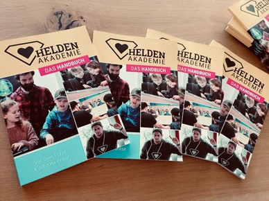 Ein Haufen Handbücher mit der Überschrift: "Das Handbuch Heldenakademie" unterlegt einer Collage aus vier Bildern mit zusammenarbeitenden Kindern.
