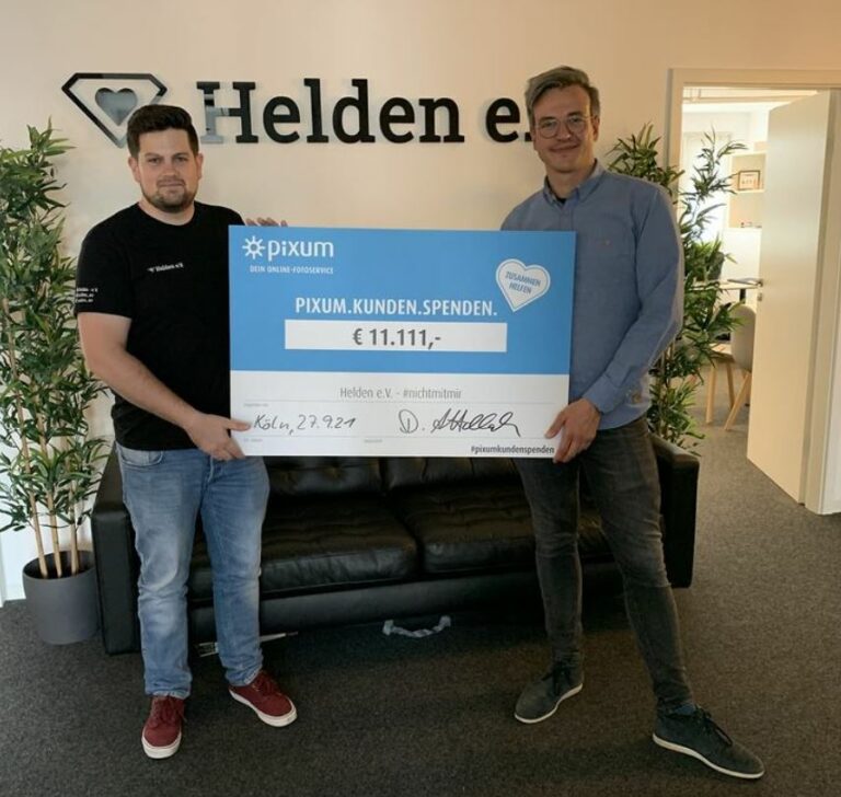 Zwei Männer halten einen Spendenschein und schauen freundlich in die Kamera. Eine Spende in Höhe von 11.111 Euro von Pixum.