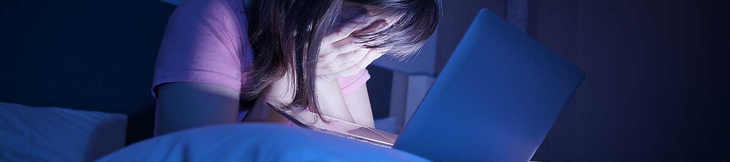 Mädchen sitzt in dunklem Zimmer auf dem Bett vor ihrem Laptop und vergräbt das Gesicht in ihren Händen