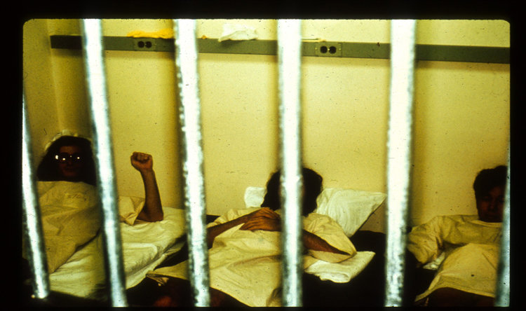 Drei Gefängnisinsassen in heller Kleidung sind in ihrer Zelle durch die Gitterstäbe zu sehen