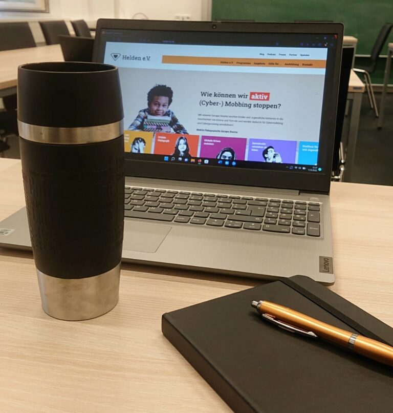 Bild eines aufgeklappten Laptops auf einem Tisch. Im Vordergrund ein Thermosbecher und ein schwarzes Notizbuch mit Kugelschreiber