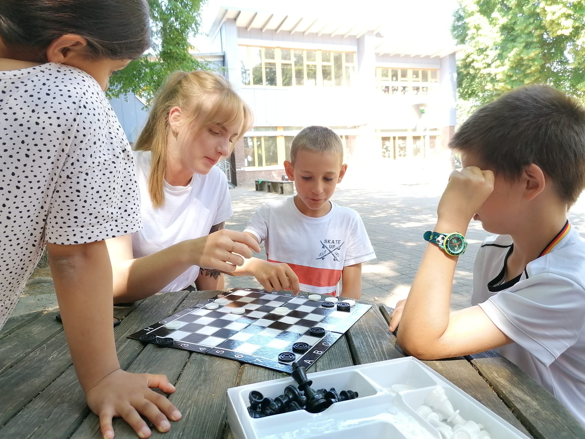 Drei Kinder und eine junge Frau sitzen an einem Holztisch auf einem Schulhof und spielen ein Brettspiel