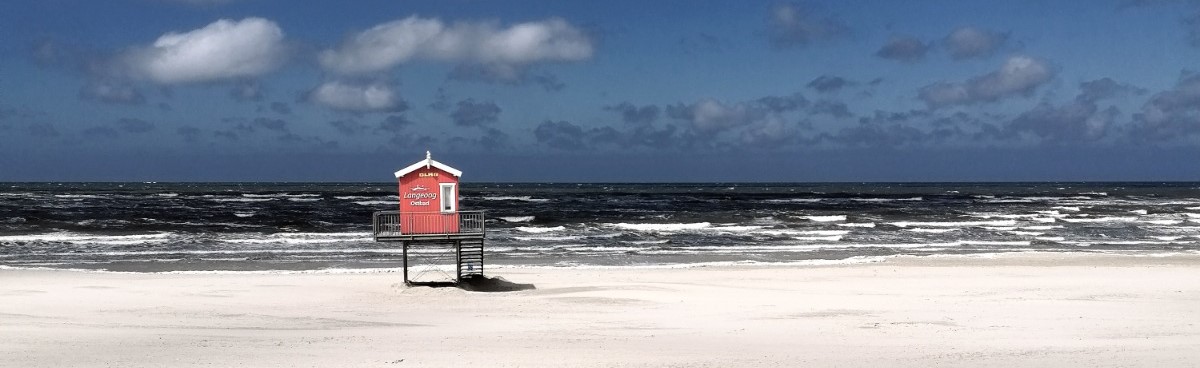 Eine kleine rote Hütte mit dem Schriftzug "Langeoog" steht an einem Strand vor dem Meer.