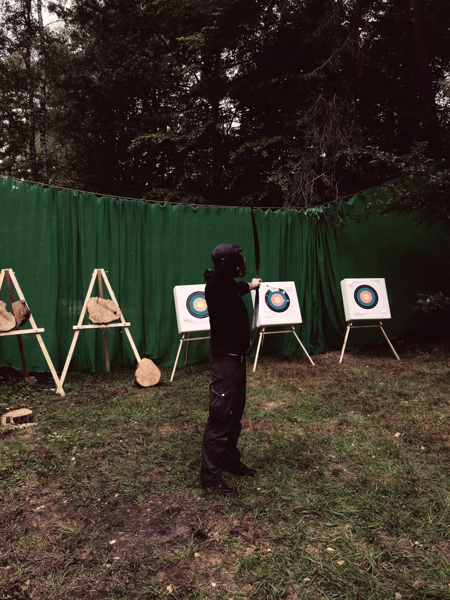 Mann in schwarzer Kleidung, den Rücken zum Betrachter gewandt, zielt mit einem Bogen auf eine Zielscheibe. Im Hintergrund sind mehrere Zielscheiben zu erkennen.