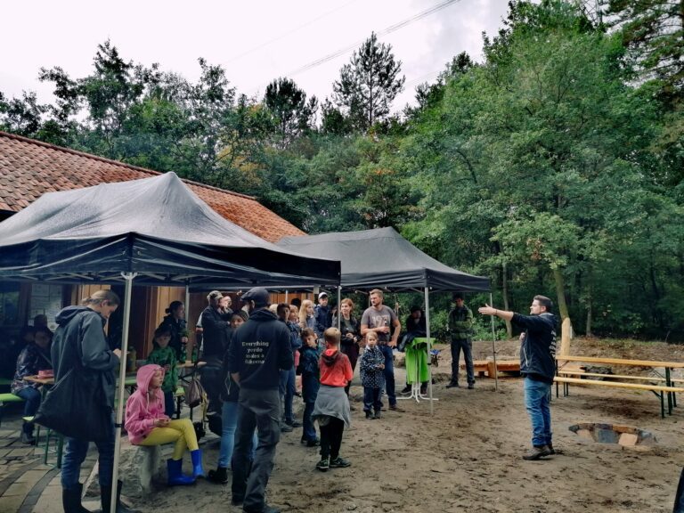Viele Menschen stehen in einer Waldlichtung vor einem Holzhaus unter zwei Pavillons verteilt. Ein Mann in schwarzem Pullover steht vor ihnen und redet.