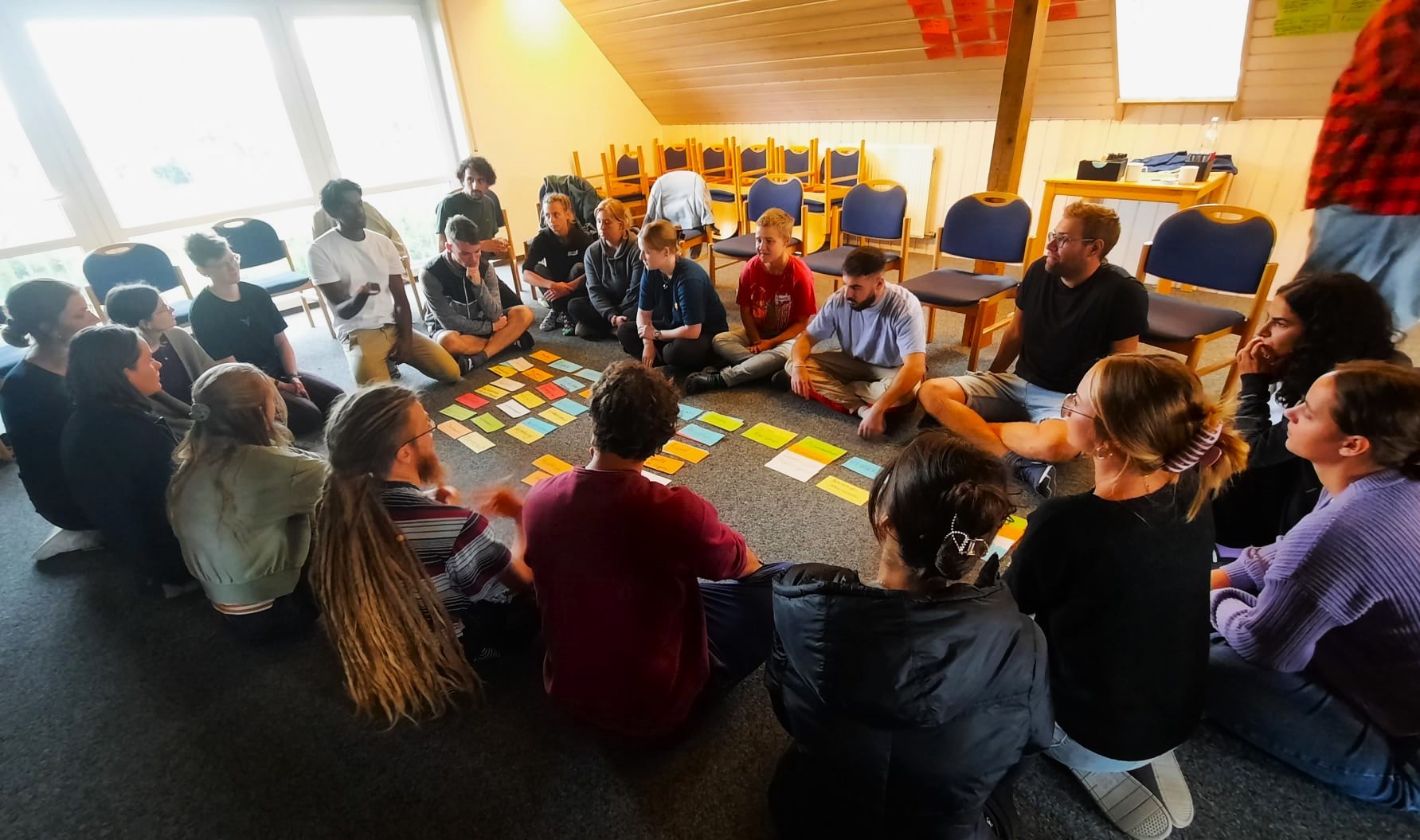 Gruppe junger Menschen die auf dem Teppichboden eines Seminarraumes im Kreis um mehrere bunte Zettel sitzen.