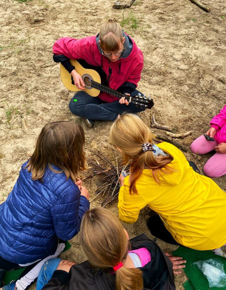 Mittig oben spielt eine junge Frau mit pinker Jacke Gitarre. Unten mittig drei Mädchen mit bunten Jacken die ein Lagerfeuer entzünden wollen.