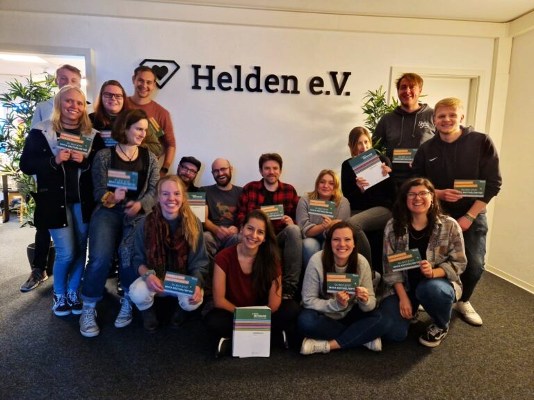 Eine Gruppe Menschen halten ein Buch hoch und lächeln in die Kamera. Im hintergrund an der Wand ein Schriftzug "Helden e.V."