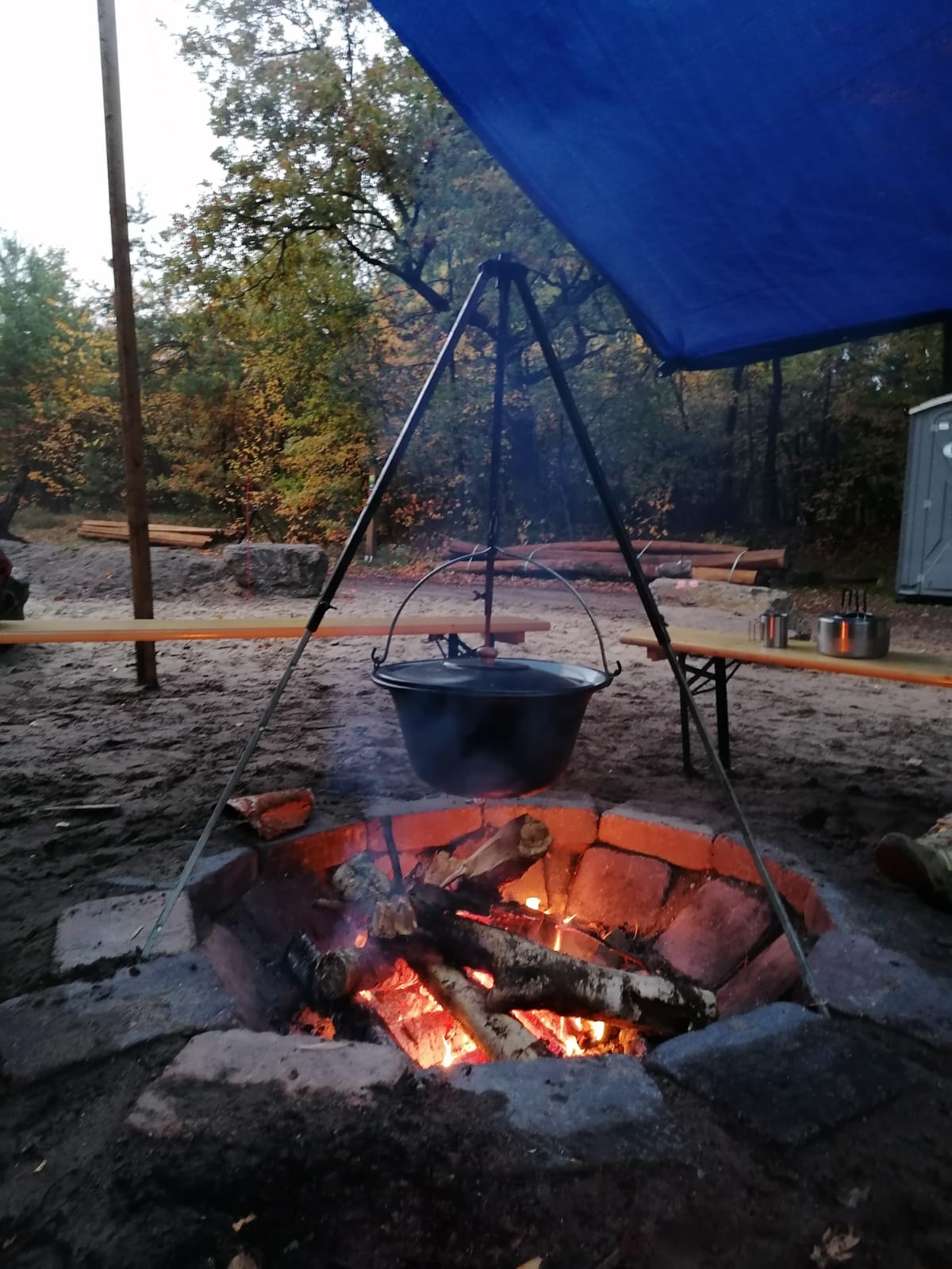 Dunkler großer Topf mit Deckel häng an einem Gerüst über einem Lagerfeuer. Im Hintergrund sind Holzbänke und rechts oben ein blauer Sonnenschutz.