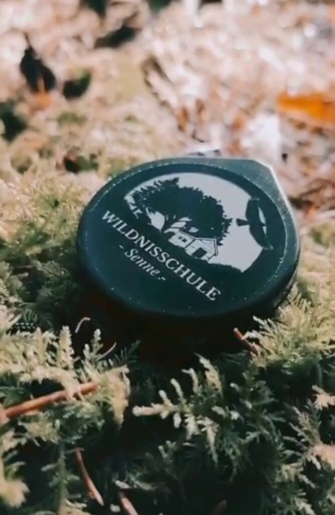Ein rundes Kunststoffstück mit einem Logo und Schriftzug "Wildnisschule Senne" liegt auf Waldboden