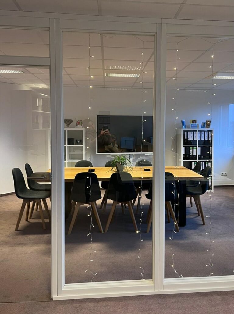 Durch ein Fenster an dem Lichterketten hängen steht ein Tisch mit schwarzen Stühlen in einem Seminarraum