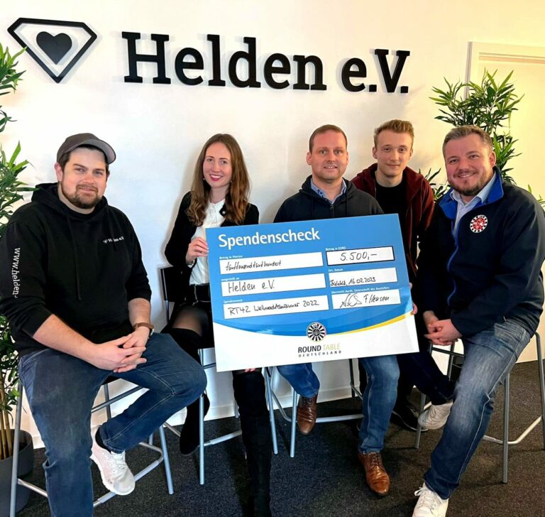 Fünf Personen sitzen auf Stühlen vor einer Wand. Zwei von ihnen halten einen großen Spendencheck in die Kamera