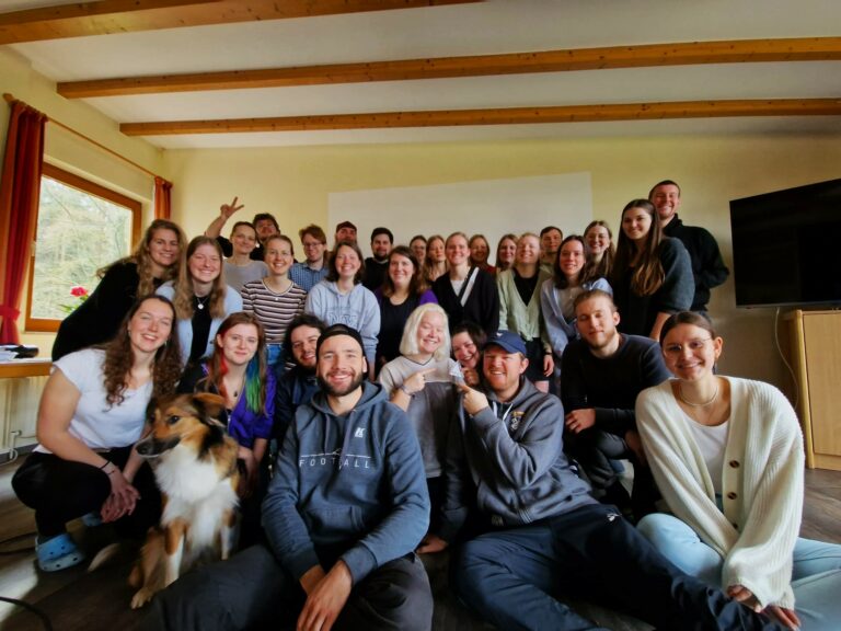 Eine Gruppe junger Menschen posiert vor einer Wand für ein Gruppenfoto und lächelt in die Kamera.