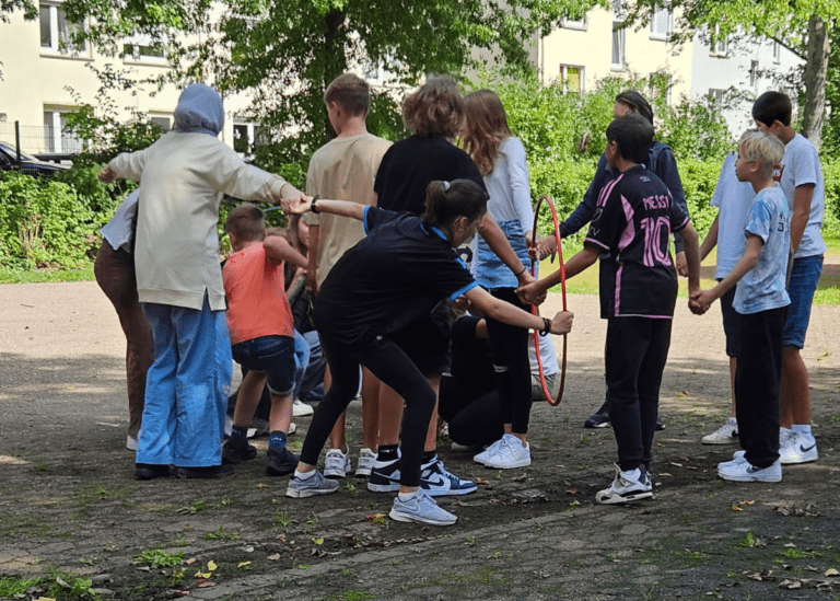 Schulklasse muss bei einer Teamaufgabe durch einen Reifen steigen. Eine Maßnahme für den Zusammenhalt und gegen soziale Isolation.